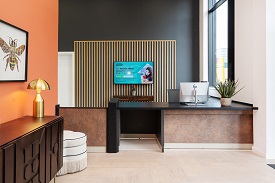 The reception desk for Zinc Quarter student accommodation. A desk with a digital screen behind  in the background and a shelving unit to the left of the photo with a lamp.